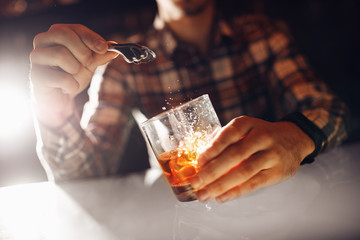 Barman puts ice pliers with splash into glass of whiskey bar. Dark brown background