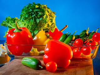 Bright vegetable still life. Fresh vegetables in a clear bowl on the table. Preparation of Greek salad. Salad of fresh vegetables. Products on a blue background. The farm's products.