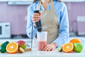 Woman in apron uses hand blender to mixing fresh fruits for prepare smoothie in the kitchen at home