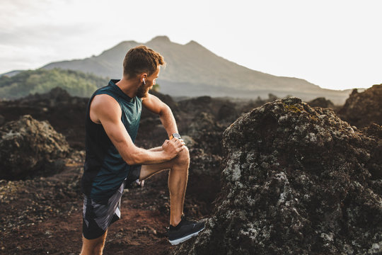 Young hipster runner with beard stretching and warming-up for trail running outdoors. Listening music in air pods. Mountain view on background.