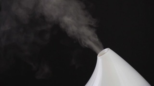 Aromatherapy. Essential oil aroma diffuser humidifier diffusing water articles in the air. Electric ultrasonic device emitting atomized water droplets against black background with copy space.