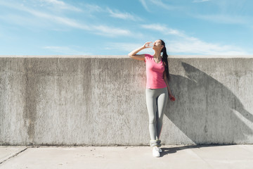 a girl in sportswear stands and drinks water from a glass against the sky and...