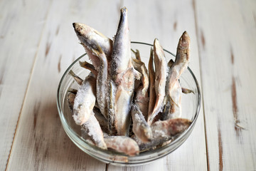 Frozen sprat herring fish in a glass bowl on a wooden light background.