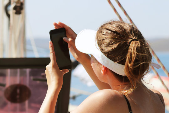A young slender girl in a swimsuit is taking pictures with a mobile phone sitting on the stern of a sailing yacht. A luxury holiday on the water for wealthy people. Prestigious lifestyle. Vacation and