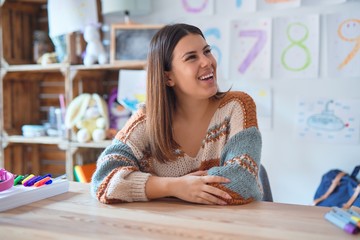 Young beautiful teacher woman wearing sweater and glasses sitting on desk at kindergarten looking...