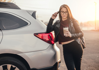 Brunette young woman with smartphone and cup of black coffee near brand new car at city parking
