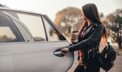Brunette young woman with smartphone and cup of black coffee near brand new car at city parking