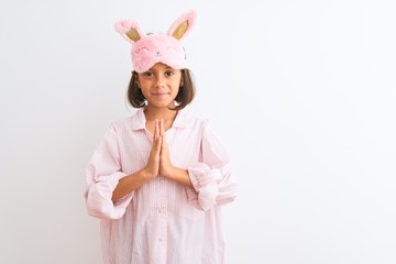 Beautiful child girl wearing sleep mask and pajama standing over isolated white background praying...