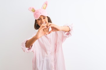 Beautiful child girl wearing sleep mask and pajama standing over isolated white background smiling in love doing heart symbol shape with hands. Romantic concept.
