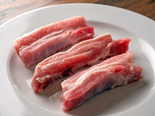 Uncooked raw pork belly slices on a white plate and wooden table.