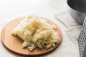 Grated white cabbage on a round wooden board on a white table. Near a metal grater and a bowl.