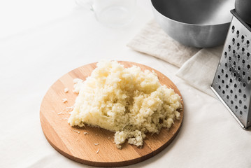 Grated white cabbage on a round wooden board on a white table. Near a metal grater and a bowl.
