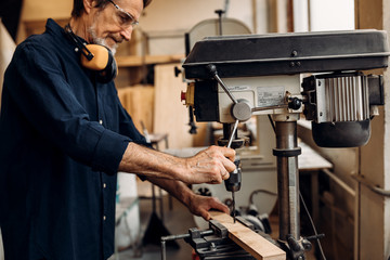 Mature worker in carpentry preparing furniture parts using a drilling machine