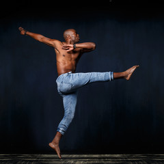 A black athletic man in jeans with a naked torso is dancing against a dark background.