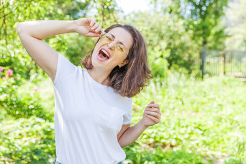 Beautiful young brunete woman with brown hair in trendy yellow sunglasses resting and smiling on park or garden green background. European woman. Portrait of happy teenage girl relaxing outdoor.