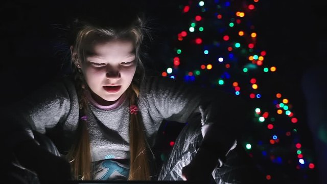 child girl watching cartoon on tablet pc at new year night. surprised funny smiling toddler kid watching Christmas movie on phone. colorful Christmas tree lights bokeh background. happy 2021 year