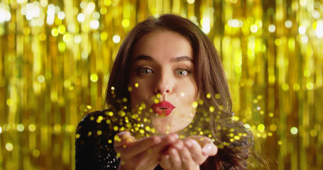 Close up of the young Caucasian beautiful brunette woman with red lips looking at the camera and blowing away golden dust from her palms, then smiling happily. New Year Eve concept. Portrait.
