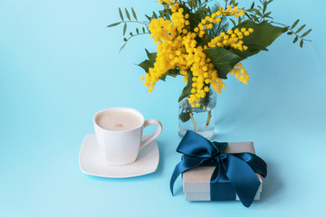 Obraz na płótnie Canvas Cup of coffee, gift in a box and yellow spring flowers on a blue background. Valentines day or womens day concept.