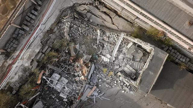 An aerial view of demolished building next to the remained construction. Concrete beams, wooden elements and lots of debris are there. Some workers in orange helmets are walking along the ruined
