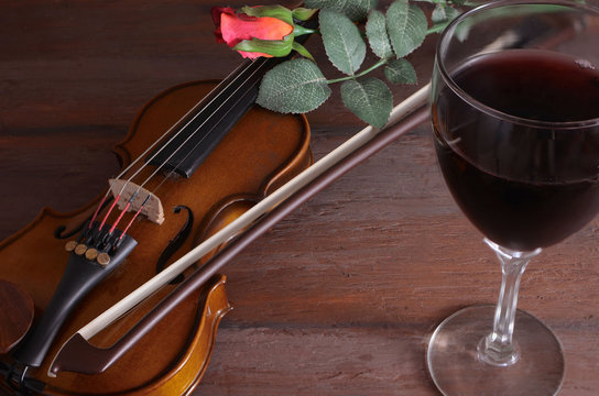 Romantic image of a violin with a glass of red wine and a single red rose on a rustic wooden table.