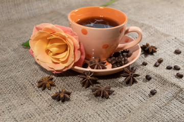 Close-up. On the table is a cup of coffee. On a saucer lies a rose. Nearby there are coffee beans and star anise.