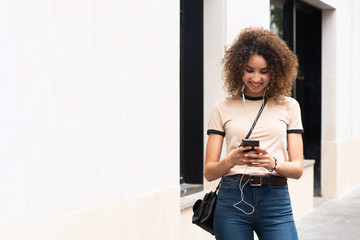 smiling young african american woman standing with cellphone and earphones in city