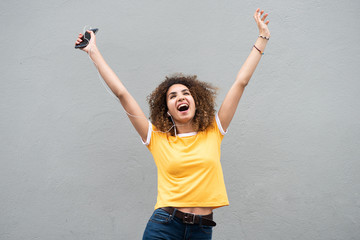happy young woman with arms raised holding mobile phone and listening to music by gray background