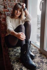 Lifestyle natural light portrait young adorable woman in white cozy sweater black boots tights sits on a windowsill and looks at the camera in grunge style loft