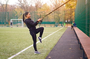 Girl athlete functional training on sportground. Mixed race young adult woman do workout with suspension system. Healthy lifestyle. Stretching outdoors playground.