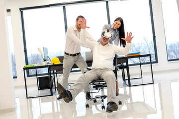 Fototapeta na wymiar Young smiling business people push senior colleague wearing virtual reality headset sitting in chair