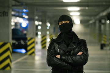 Criminal in black leather jacket and balaclava on head crossing arms by chest - obrazy, fototapety, plakaty