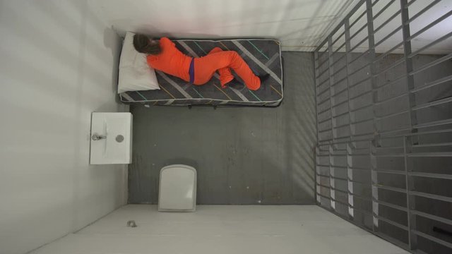 Female Prisoner in Prison Cell. The woman is trying to sleep in bed. Aerial view from above. Dressed in Orange