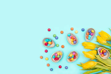 Easter border composition with chocolate eggs with colorful candies bonbons, yellow tulips on blue background. Stylish decor minimal concept. Copy space. Festive flat lay greeting card