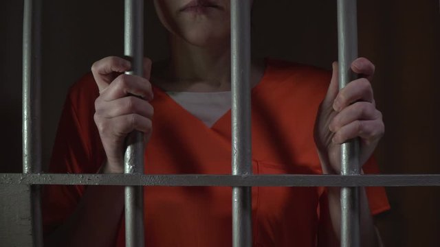 Female Prisoner / Inmate behind bars in a Jail / Prison Cell. The criminal is holding the bars. Tracking shot back. Orange