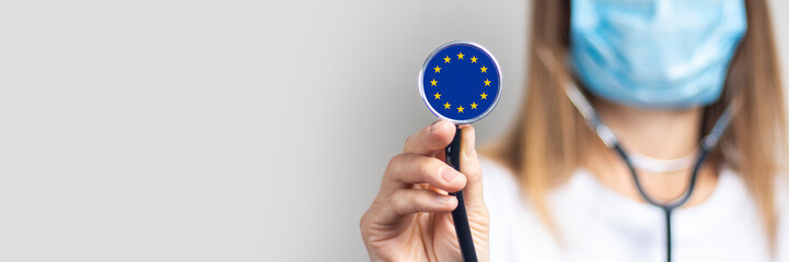 Young woman in a medical mask holds a stethoscope with the flag of European Union on a light...