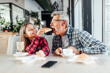 Cute stylish granddaughter wearing red shirt feeding grandfather croissant, having breakfast together.