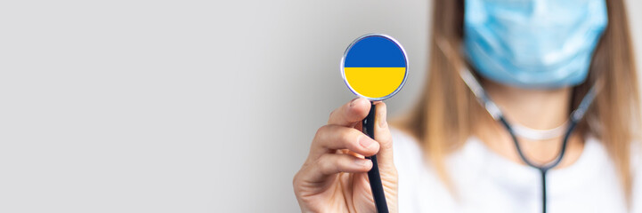 Young woman in a medical mask holds a stethoscope with the flag of Ukraine on a light background....