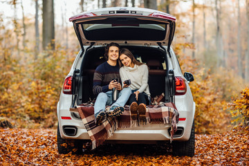 Beautiful pretty couple enjoying picnic time on the forest. They holding cup with tea and sitting in a car.