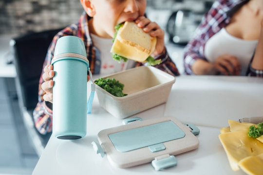 Cropped image, small boy eating his sandwich before school at home kitchen, thermos on his hands.
