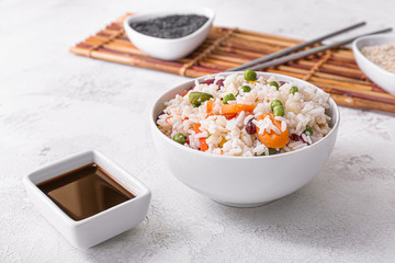 Boiled rice with vegetables in bowl and sauce on white background