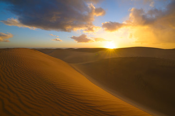 Obraz na płótnie Canvas Sunset in the desert, sun and sun rays, blue sky and Beautiful clouds. Golden sand dunes in desert in Maspalomas, Gran Canaria at Canary islands, Spain