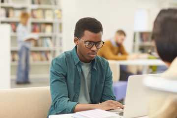 Portrait of African teenage boy studying in college library, using laptop with other students in background, copy space