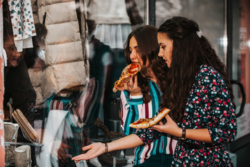 Portrait of two beautiful young women talking and eating pizza outdoors, and pointing finger in the shop window