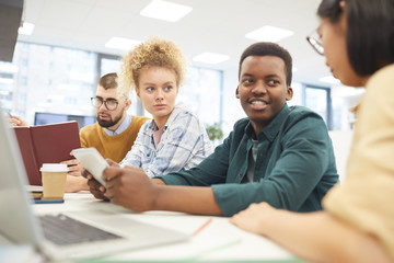 Multi-ethnic group of students studying in college library, focus on African-American man smiling at partners