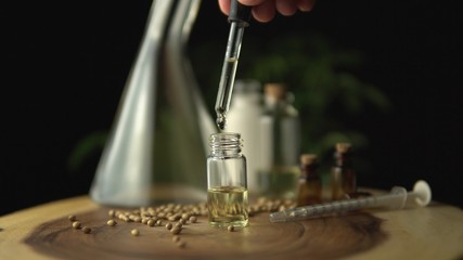 Macro close up of droplet pipette dosing biological and ecological oil of the hemp plant.  Pharmaceutical herbal cbd oil in the small jar on the wood circle tree ring table. Organic pharma concept.