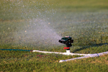 Up close photo of an automatic impact water sprinkler mounted on a custom base of white PVC pipes watering green grass. Water sprays from a sprinkler