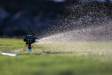 Water sprays from an automatic impact water sprinkler mounted on a custom base of white PVC pipes watering green grass.