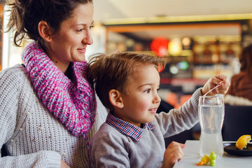 Small caucasian boy side view three years old mother and son young woman sitting by the table at the restaurant or cafe child holding plastic spoon or straw while preparing drink having fun