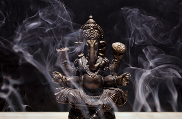 Hindu god Ganesha on black background. Statue on wooden table with a smoke of incense. 