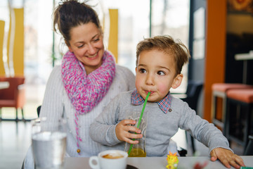 Portrait of small three years caucasian boy kid child sitting by the table with his mother young woman female at cafe having a juice or soda drink holding a glass with a straw drinking